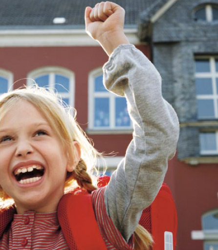 Das Bild zeigt ein lachendes Mädchen mit Schultournister vor einer Schule